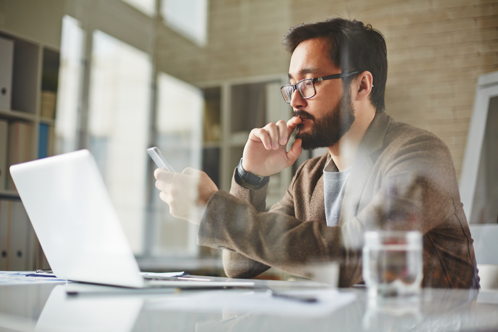 Man in office in front of laptop using his mobile phone